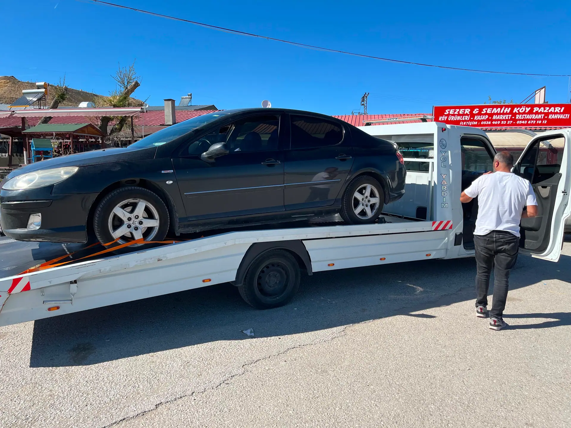 Köy pazarı önünde çekici platformunda taşınan siyah sedan otomobil ve aracın yanındaki görevli